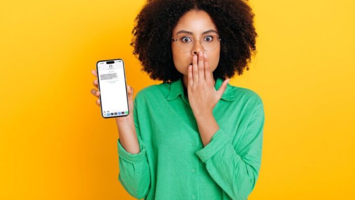 Black woman in a green shirt holding an iPhone looking surprised. Yellow background.