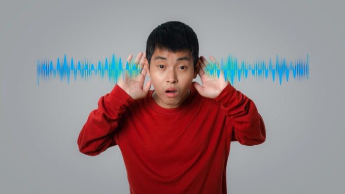 a man with short dark hair wearing a long-sleeved red shirt has his hands up to his ears as if to amplify the sound. blue angular sound waves are superimposed over the image