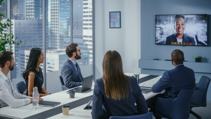 a conference room of people around a desk looking at the tv screen with another meeting participant on screen