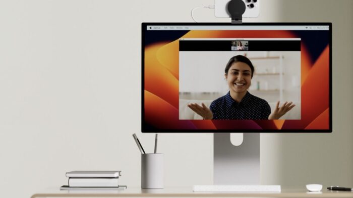 desk with a computer monitor on it. monitor has an iPhone attached to the top. on screen is a woman smiling with her palms facing up on each side of her