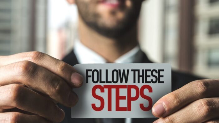 Small white sign that says "FOLLOW THESE STEPS." Follow these is black. Steps is red and larger. Sign is being held by a man's hands. In the background is a blurry image of the mouth, chin, and upper chest of a man dressed in a suit.