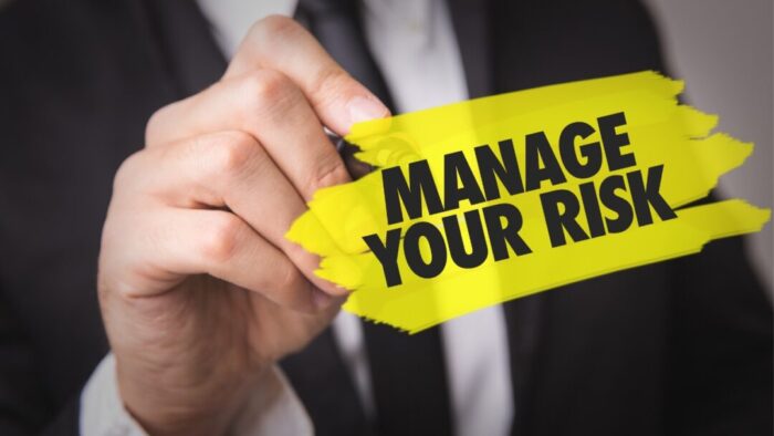 MANAGE YOUR RISK written in foreground with yellow highlighter behind it. Behind that is a blurred image of a person in a black suit, white shirt, and black tie with hand outstretched holding the highlighter.