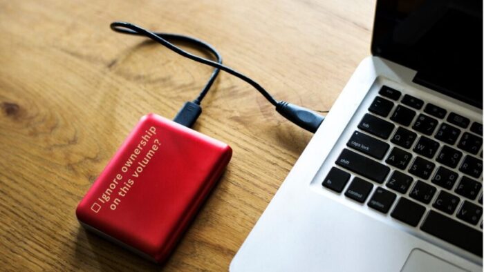 Left side of a Mac laptop on a wooden desk. Red hard drive is plugged into it. White text overlay on the hard drive reads [checkbox] Ignore ownership on this volume?