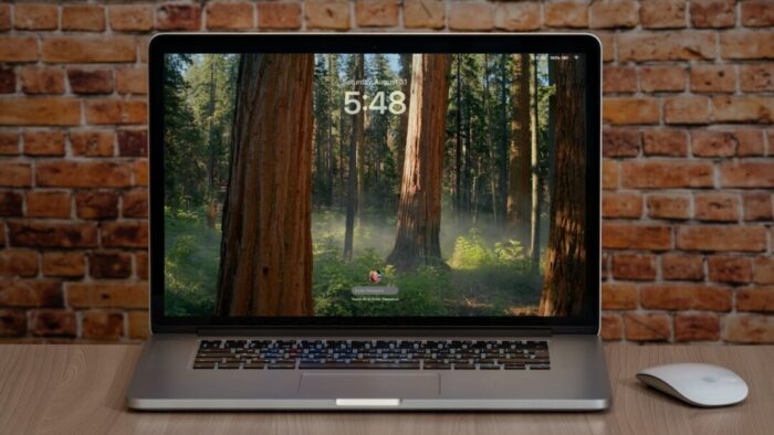 MacBook at the Lock Screen with a forest as the screen's background. Mac is on a wooden table against a brick wall. Computer mouse is to the right of the Mac