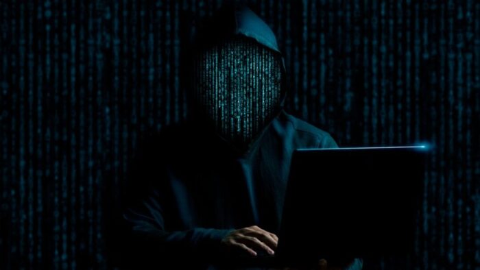 nearly all black image with the silhouette of a person in a hoodie with hands in the direction of a laptop keyboard off to the right. faint binary code across the obscured face of the person.