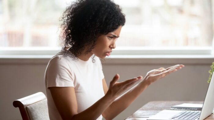 side view of a woman with her palms turned upwards in an expression of frustration. she's sitting at a desk looking at a laptop computer. there's a large bright window in the background.