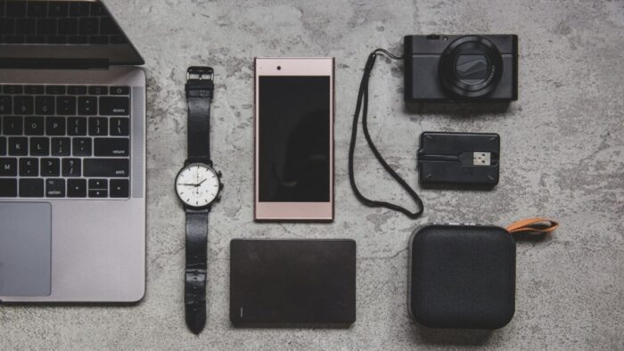 from left to right, partial Mac laptop, an analog watch, cellphone, hard drive, digital camera, and battery pack.