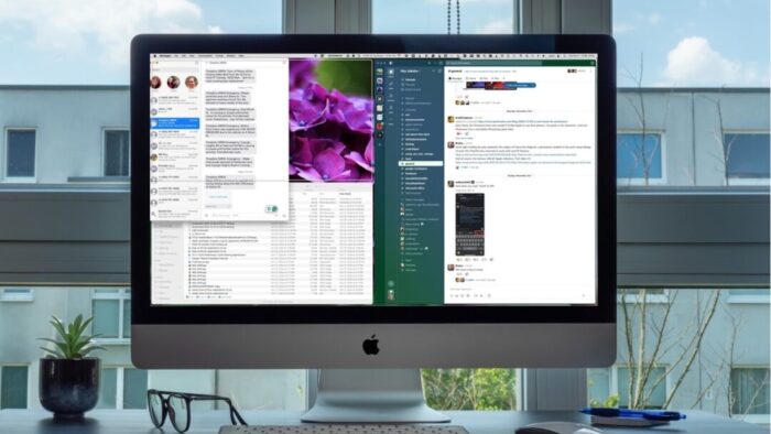 looking at an iMac that's in front of a window looking out to the next building over with a partly cloudy sky at the top. a small plant, pair of glasses, keyboard, mouse, and pen all sit on the desk below the iMac screen.