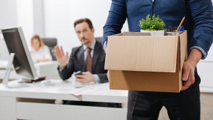 person in a blue sweater carrying a cardboard box of personal items as they leave an office. two other employees are blurred in the background.