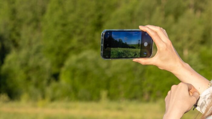 a person's right hand lifting up an iPhone to take a picture with the Camera app. Background is a green nature seen with trees.