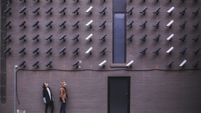 two people standing against a painted brick wall wearing sunglasses and looking up at the large grid of security cameras pointed at them