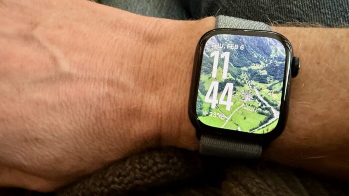 picture of an Apple Watch on a person's right wrist. watch face is of a verdant green town backed up to a mountain range