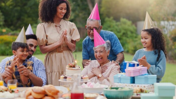 three generations of a Black family at an outdoor birthday party. everyone is wearing a pointed hat. gifts and food are abundant in front of them.