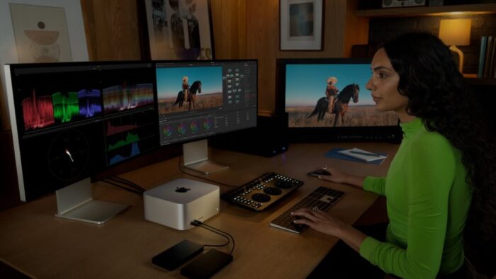 dimly lit room with a woman in a green shirt sitting in front of three large monitors and a Mac Studio computer in the middle, along with two keyboards and a mouse.