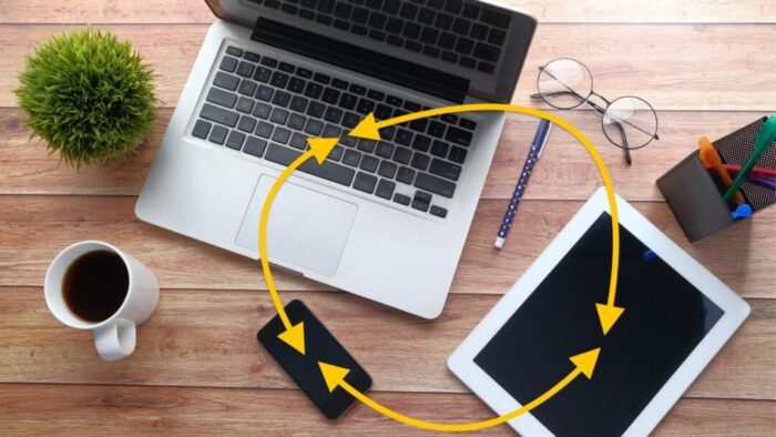 overhead view of a desk with a laptop, iPad, and iPhone on it. superimposed over the devices are yellow circular-ish lines with arrows on both ends, indicating connections between each device.