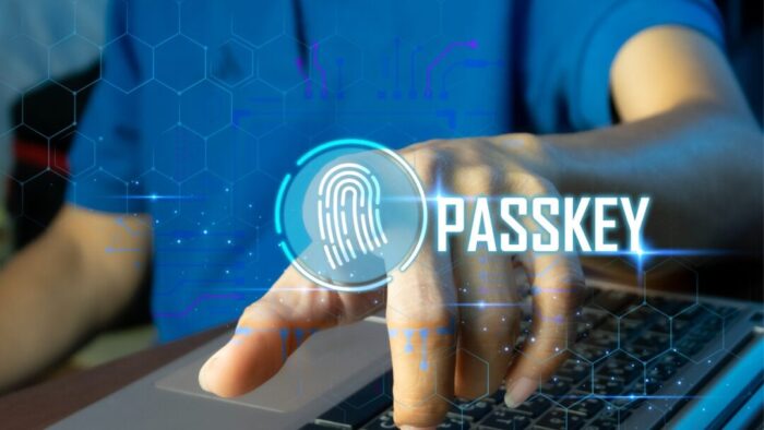 torso of a person in a blue shirt with their hand on a laptop keyboard. index finger is pointing straight out. overlaid the finger is a fingerprint icon and the word passkey
