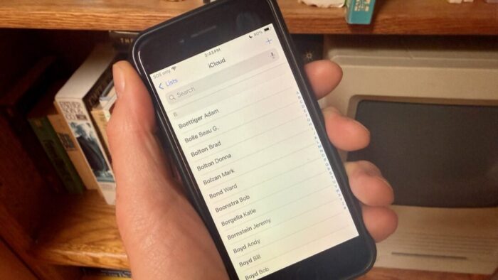 a disembodied left hand holding an iPhone that is showing a list of contact names