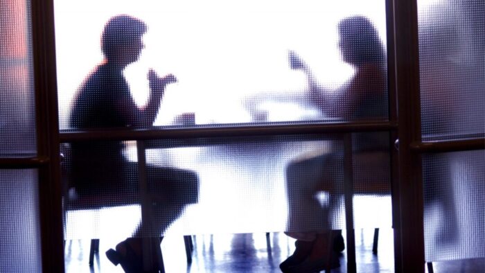 photo of two people sitting at a table talking. the image is blurred because they are behind a frosted glass wall.