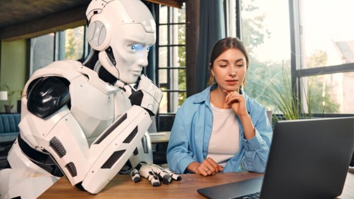 a larger than life white android robot leaning in while looking at an open computer with a woman sitting next to it.