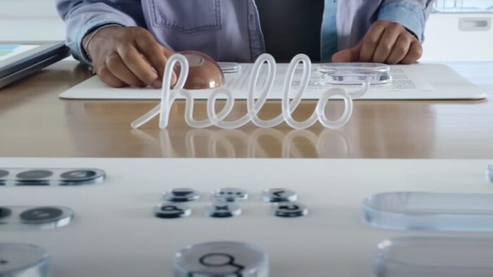 screenshot of the hands of a person resting on a table. "hello" looks like a clear white neon sign standing up in front. in the foreground, there are glass beads on a white surface. you can faintly see shapes being reflected from under the glass beads