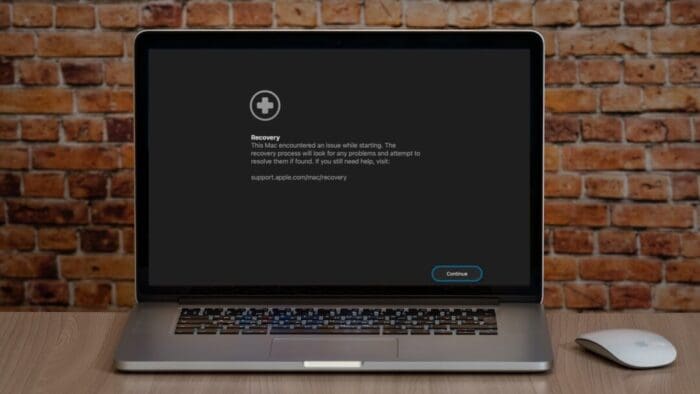 Mac laptop in recovery mode. Mac is on a table with a brick wall background. Magic Mouse is to the right of the Mac.