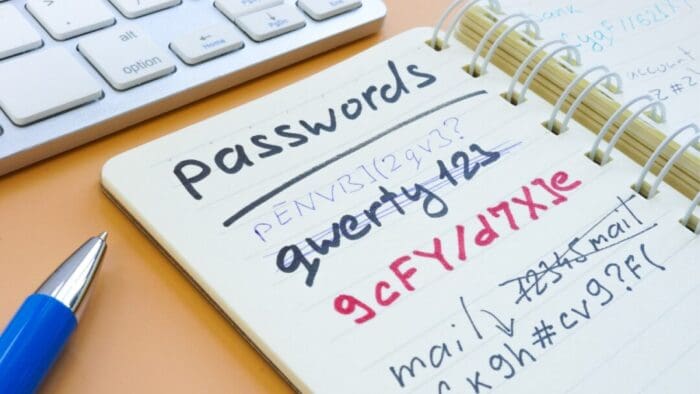 small open spiral notebook with "passwords" at the top of the page and several passwords (some crossed out) listed on the page. to the left is the tip of a pen. above the notebook is the corner of a keyboard
