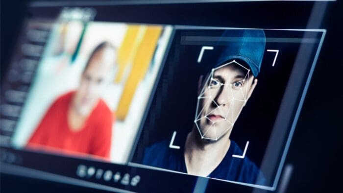 AI-generated image. side view of a computer screen with two faces on it. The face farther away is blurred, but full of color. the face closer up is in focus, but has a grid on it indicating it's calculating facial features.