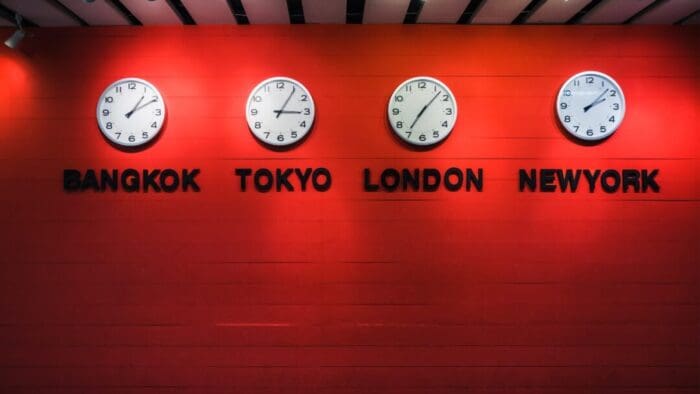 dimly lit deep red wall with four analog clocks on it, each set to a different time according to the city named below them. The cities named are Bangkok, Tokyo, London, and New York.