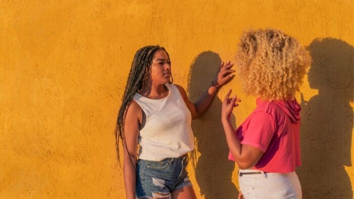 two Black women facing each other against a deep yellow wall.