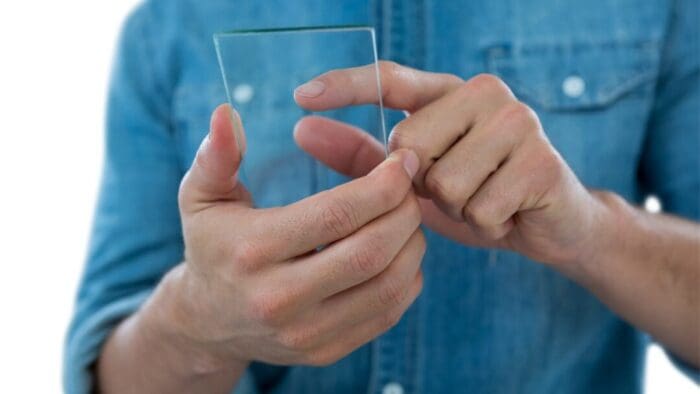 torso of a man in a blue denim shirt holding a slab of glass like a phone while tapping on the glass.
