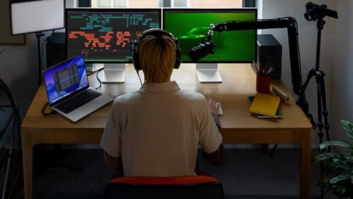 standing behind a guy at his desk. the desk has a media recording set up with two monitors, a bright light (turned off), Mac, and a microphone.