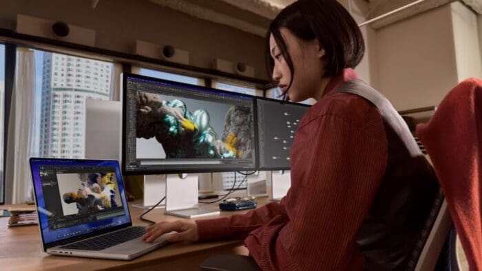 a woman sitting at a desk facing a window. there are two large monitors and one laptop on the desk. she is working at the laptop.
