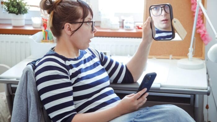 woman with her hair up in a bun is reclining in a chair holding her phone in one hand and a mirror in the other. She's looking into the mirror with a surprised look on her face