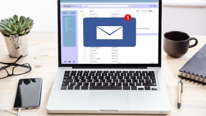 an open Mac laptop on a desk with a few accessories. on the screen is email with a Microsoft Outlook icon in the middle