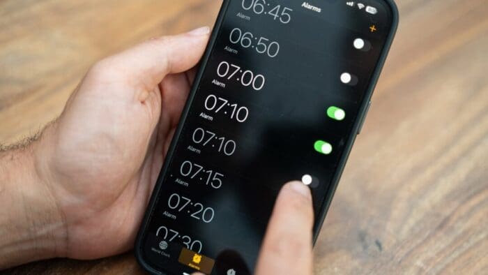 a person holding an iPhone in their left hand and using the phone with their right. Screen shows a full screen of alarms from 06:45 to 07:30. This person must need a lot of reminders to get out of bed in the morning!